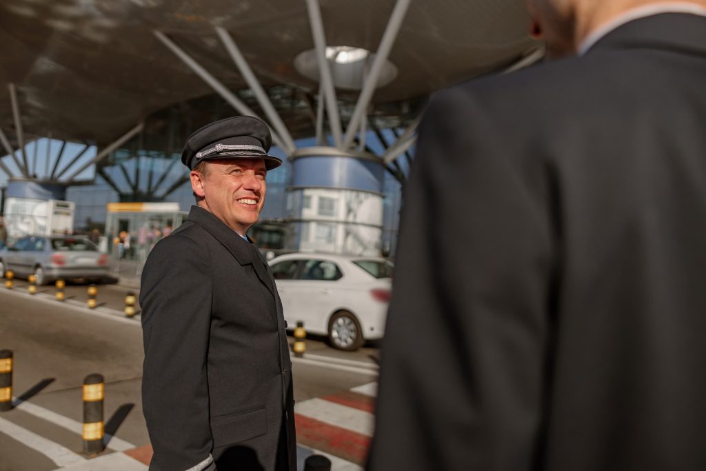 A smiling chauffeur in uniform talks to a colleague outside an airport terminal, with cars and modern architecture in the background.